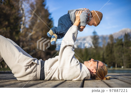 Happy family. Young mother playing with her baby boy infant oudoors on sunny autumn day. Portrait of mom and little son on wooden platform by lake. Positive human emotions, feelings, joy 97887221