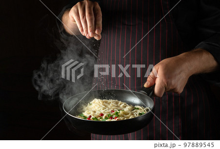 The cook adds salt to the spaghetti in a steaming hot pan. Recipe or menu for restaurant or hotel on black background. The cook adds salt to the spaghetti in a steaming hot pan. Recipe or menu for restaurant or hotel on black background. 97889545