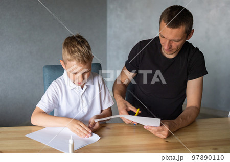 A cute boy is sitting with his dad at the table and collecting origami. Cutting a white sheet of paper A cute boy is sitting with his dad at the table and collecting origami. Cutting a white sheet of paper 97890110