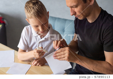 A cute boy is sitting with his dad at the table and collecting origami. Holds a white sheet of paper A cute boy is sitting with his dad at the table and collecting origami. Holds a white sheet of paper 97890113