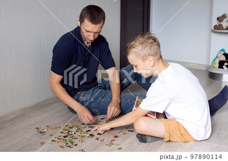 A cute boy with a cheerful dad is collecting a puzzle lying on the floor in a room A cute boy with a cheerful dad is collecting a puzzle lying on the floor in a room 97890114