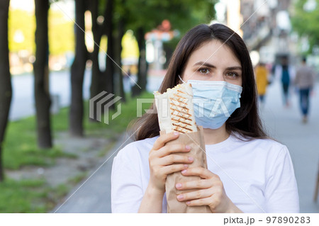 A masked deliverywoman holds an order - fast food, street food and home delivery by courier during a pandemic. 97890283