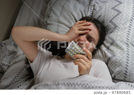 A tired sleepy woman lies in bed, holding her head with her hand, pills in her hands. 97890685