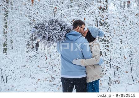 a loving couple in the winter forest is kissing. a man holds a Christmas tree on his shoulder a loving couple in the winter forest is kissing. a man holds a Christmas tree on his shoulder 97890919