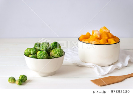 Frozen vegetables assortment. Frozen Brussels sprouts and pieces of pumpkin in white bowls on white table Frozen vegetables assortment. Frozen Brussels sprouts and pieces of pumpkin in white bowls on white table 97892180