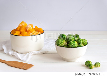 Frozen vegetables assortment. Frozen Brussels sprouts and pieces of pumpkin in white bowls on white table Frozen vegetables assortment. Frozen Brussels sprouts and pieces of pumpkin in white bowls on white table 97892182