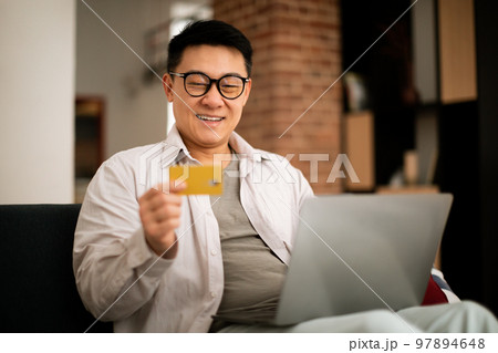 E-commerce concept. Happy asian mature man holding credit card and shopping online with laptop, sitting on couch at home 97894648