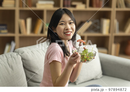 Healthy vegan diet concept. Happy asian lady sitting on sofa at home with bowl of fresh vegetable salad Healthy vegan diet concept. Happy asian lady sitting on sofa at home with bowl of fresh vegetable salad 97894872