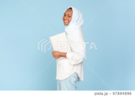 Cheerful black muslim woman holding laptop computer posing over blue studio background, free space 97894896