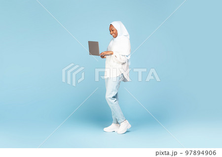 Positive african american muslim woman in hijab posing with laptop over blue studio background, full length shot 97894906