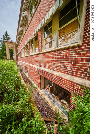 Detail of exterior wall of abandoned brick building with broken glass and overgrown fields Detail of exterior wall of abandoned brick building with broken glass and overgrown fields 97894935