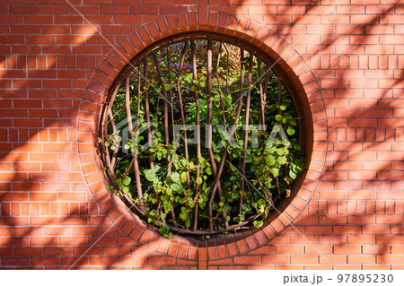 Brick wall with circle opening blocked by abstract steel bars and filled with greenery in center straight on Brick wall with circle opening blocked by abstract steel bars and filled with greenery in center straight on 97895230