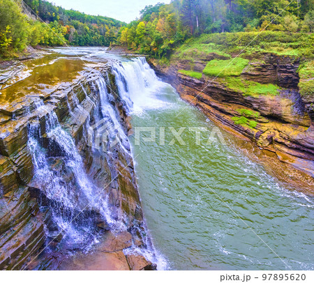 Aerial along stunning waterfall pouring over cliffs with lush forests in background Aerial along stunning waterfall pouring over cliffs with lush forests in background 97895620