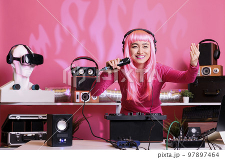 Asian musician sitting at dj table performing techno music using professional turntables, wearing headset and talking with fans using microphone. Artist with pink hair playing electronic song at night Asian musician sitting at dj table performing techno music using professional turntables, wearing headset and talking with fans using microphone. Artist with pink hair playing electronic song at night 97897464