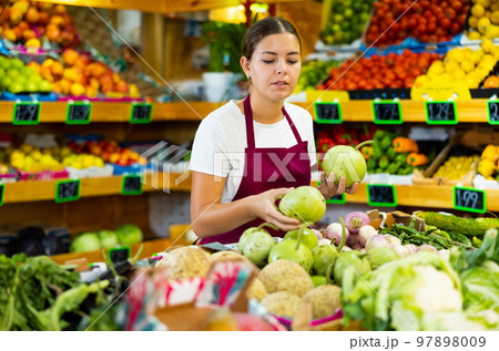 Focused young woman laying out small green pumpkins in supermarket 97898009