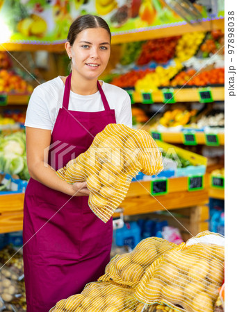 Smiling young female supermarket worker holding raw potatoes in grid Smiling young female supermarket worker holding raw potatoes in grid 97898038