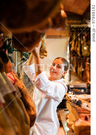 Saleswoman working behind counter in grocery shop, checking quality of jamon 97898192