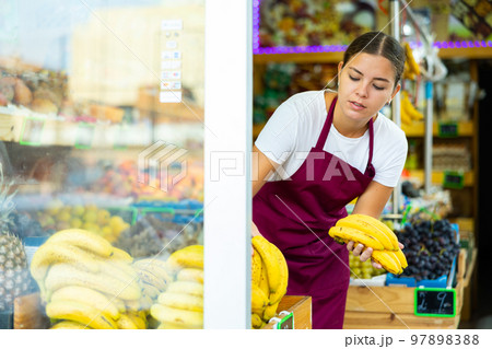 Young girl employees in uniform lay out and selling banana in grocery shop Young girl employees in uniform lay out and selling banana in grocery shop 97898388