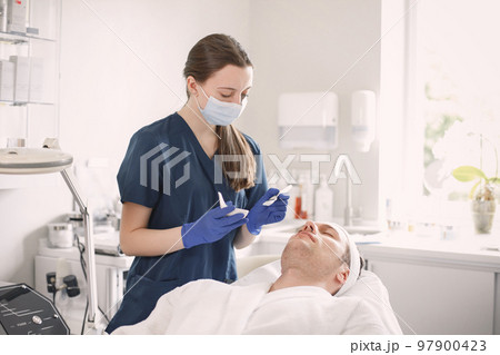 Man laying in cosmetologist cabinet and has a procedure for face skin. Female cosmetologist wearing blue medical costume and face mask. Woman applying a mask on man's face. 97900423