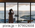 ホテルから見える、海に沈む夕日を撮影する女性 97902608