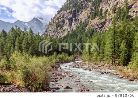 Water stream along rocky rapids in gorge between Italian Alps partly with snow-covered peaks 97910756