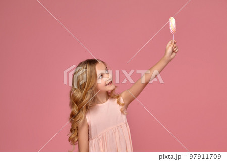 Little girl with a blond curly hair, in a pink dress is posing with a candy 97911709