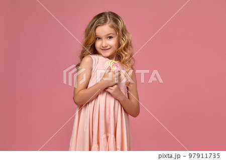Little girl with a blond curly hair, in a pink dress is posing with a candy 97911735