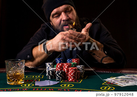 Bearded man with cigar and glass sitting at poker table in a casino. Gambling, playing cards and roulette. 97912048