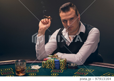 Wealthy man is smoking a cigar and playing poker with an excitement at a casino on black background. Wealthy man is smoking a cigar and playing poker with an excitement at a casino on black background. 97913164