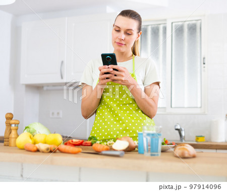 Young housewife using phone to find recipe while cooking dinner 97914096