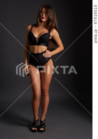 Young sexy slim tanned woman in black underwear posing against dark grey background. Young sexy slim tanned woman in black underwear posing against dark grey background. 97916010