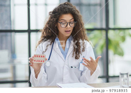 Young pharmacist woman holding pills while sitting at the table in the office Young pharmacist woman holding pills while sitting at the table in the office 97918935