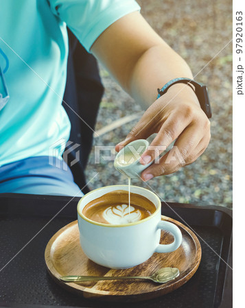 Hand of young woman pouring condensed milk into hot cappuccino on cafe table. Pouring milk in to hot coffee cup. Adding a subtle sweetness to favorite cup of coffee is delicious. Selective focus. Hand of young woman pouring condensed milk into hot cappuccino on cafe table. Pouring milk in to hot coffee cup. Adding a subtle sweetness to favorite cup of coffee is delicious. Selective focus. 97920163