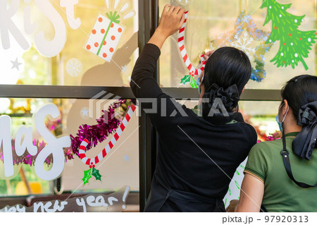 Asian women decorate beautiful stickers on window glass in front of cafe to welcome Christmas and New Year. Prepare to decorate the storefront to be beautiful during the important holidays of year. 97920313