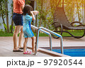 Adorable daughter in blue swimming suit going down swimming pool ladder to swim with clear blue water and her mother to take care of closely. Little girl at pool. Happy family relationship at pool. 97920544