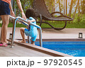 Adorable daughter in blue swimming suit going down swimming pool ladder to swim with clear blue water and her mother to take care of closely. Little girl at pool. Happy family relationship at pool. 97920545