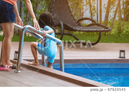 Adorable daughter in blue swimming suit going down swimming pool ladder to swim with clear blue water and her mother to take care of closely. Little girl at pool. Happy family relationship at pool. Adorable daughter in blue swimming suit going down swimming pool ladder to swim with clear blue water and her mother to take care of closely. Little girl at pool. Happy family relationship at pool. 97920545