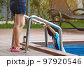 Mother wear short jeans take care her lovely daughter in blue swimming suit going down swimming pool ladder to swim with clear blue water. Mom and daughter at pool. Happy family relationship at pool. 97920546
