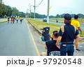 Back view of Asian sport photographer with camera sit to take picture marathon runners in running event marathon run on city street with blurred image of runners in background. Sports and photography. 97920548