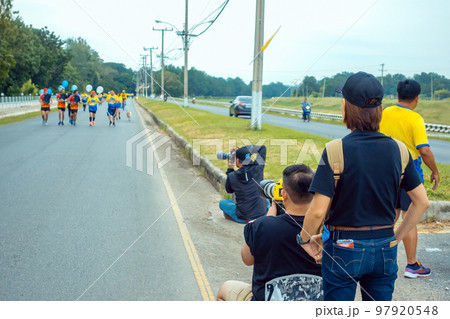 Back view of Asian sport photographer with camera sit to take picture marathon runners in running event marathon run on city street with blurred image of runners in background. Sports and photography. Back view of Asian sport photographer with camera sit to take picture marathon runners in running event marathon run on city street with blurred image of runners in background. Sports and photography. 97920548