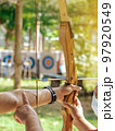 Female teacher teaches student to aim at goal. An archer teaching young man archery on field. Instructor teaching man to use bow and arrow on archery training. Archery course. Selective focus on hand. 97920549