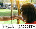 Female teacher teaches student to aim at goal. An archer teaching young man archery on field. Instructor teaching man to use bow and arrow on archery training. Archery course. Selective focus on hand. 97920550