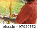 Female teacher teaches student to aim at goal. An archer teaching young man archery on field. Instructor teaching man to use bow and arrow on archery training. Archery course. Selective focus on hand. 97920552