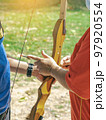 Female teacher teaches student to aim at goal. An archer teaching young man archery on field. Instructor teaching man to use bow and arrow on archery training. Archery course. Selective focus on hand. 97920554