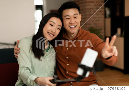 Excited korean couple middle aged man and young woman taking selfie with selfie stick, spending time together at home Excited korean couple middle aged man and young woman taking selfie with selfie stick, spending time together at home 97928102