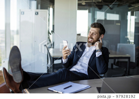 Happy middle aged businessman relaxing in office, resting feet on table using cellphone and listening to music Happy middle aged businessman relaxing in office, resting feet on table using cellphone and listening to music 97928192