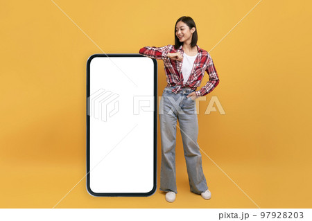 Happy japanese lady leaning on big smartphone with blank white screen and smiling, yellow studio background, mockup Happy japanese lady leaning on big smartphone with blank white screen and smiling, yellow studio background, mockup 97928203