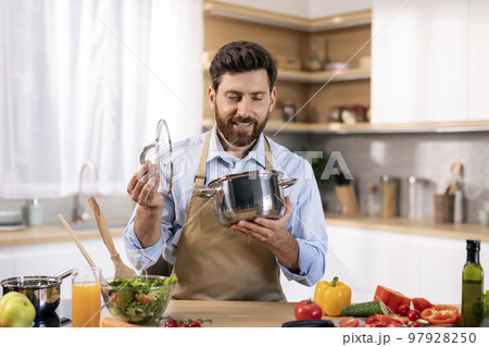 Happy millennial caucasian bearded male chef in apron prepare food, looks at pan at table Happy millennial caucasian bearded male chef in apron prepare food, looks at pan at table 97928250