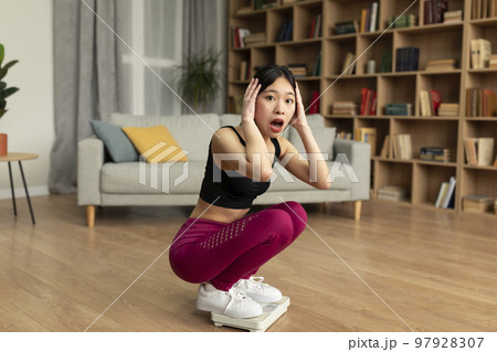 Shocked asian lady sitting on scales, terrified of weight gain at home, touching head and looking at camera, free space Shocked asian lady sitting on scales, terrified of weight gain at home, touching head and looking at camera, free space 97928307