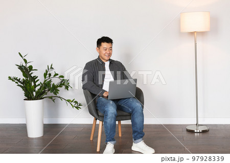 Cheerful middle aged korean man typing on laptop surfing in internet, chatting in living room Cheerful middle aged korean man typing on laptop surfing in internet, chatting in living room 97928339